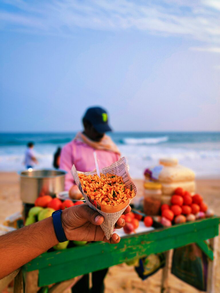 pexels-photo-10835609-10835609 Hand holding a cone of bhel puri in front of a beachside snack vendor with ocean in background.