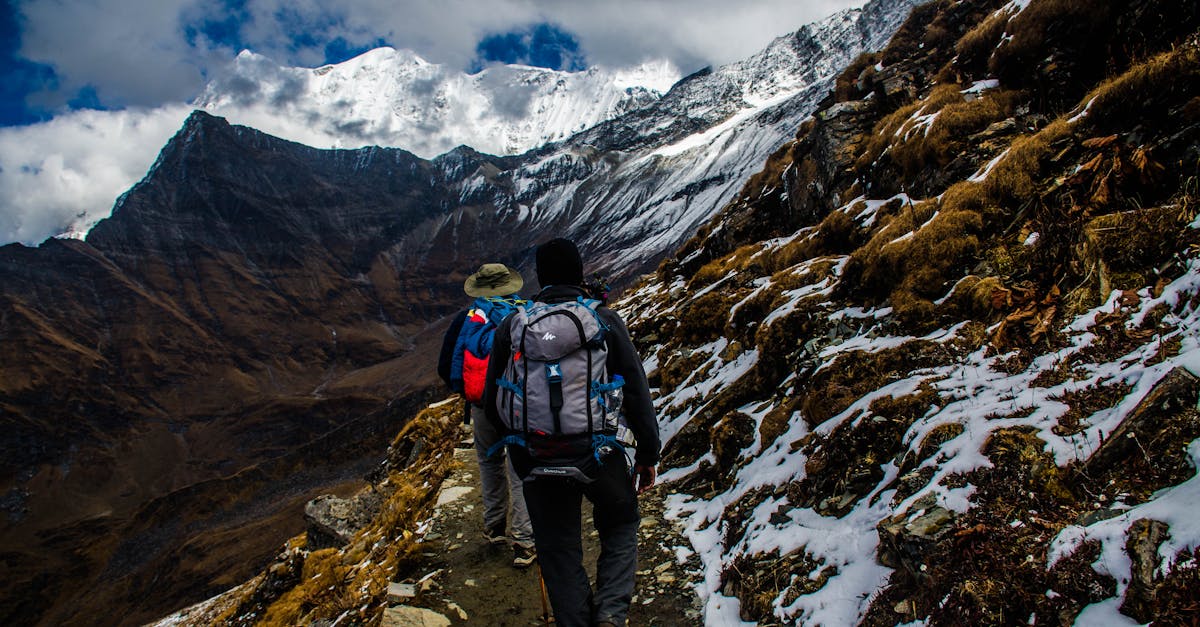 Two hikers navigating a snowy trail in rugged mountainous terrain under dramatic skies.