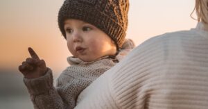 A mother carries her bundled-up toddler at a beach during a picturesque winter sunset. सर्दियों में बच्चों की त्वचा और स्वास्थ्य की देखभाल: रूखेपन से बचाव और इम्युनिटी बढ़ाने के 10 ज़रूरी उपाय