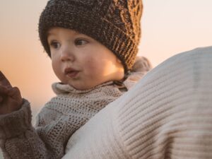 A mother carries her bundled-up toddler at a beach during a picturesque winter sunset. सर्दियों में बच्चों की त्वचा और स्वास्थ्य की देखभाल: रूखेपन से बचाव और इम्युनिटी बढ़ाने के 10 ज़रूरी उपाय