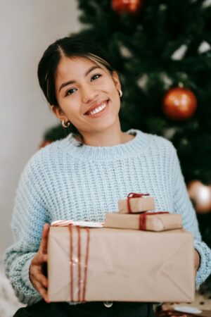 Content young ethnic female with present boxes looking at camera against fir tree during New Year holiday in house क्रिसमस उपहार और 'जीरो-वेस्ट' दर्शन: खुशियाँ बाँटने का एक नया और टिकाऊ तरीका