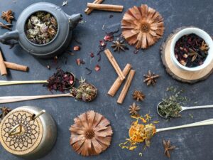 A flat lay of various herbal tea ingredients, spices, and rustic utensils on dark surface.
