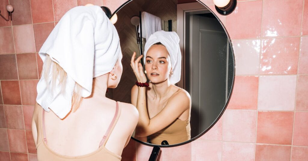 A young woman with a towel in a pink bathroom, reflecting in round mirror as she performs skincare.