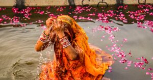A woman performing Chhath Puja in traditional attire amidst vibrant floral decorations by a river. मकर संक्रांति 2026: जानें स्नान-दान का महामुहूर्त, सूर्य देव की विशेष पूजा विधि और महत्व