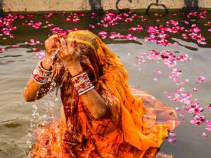 A woman performing Chhath Puja in traditional attire amidst vibrant floral decorations by a river. मकर संक्रांति 2026: जानें स्नान-दान का महामुहूर्त, सूर्य देव की विशेष पूजा विधि और महत्व