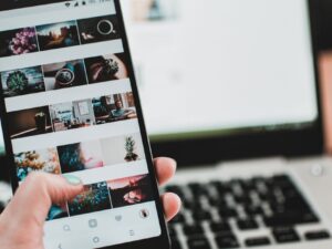 A person uses a smartphone to browse social media in front of a laptop, emphasizing modern technology and lifestyle.