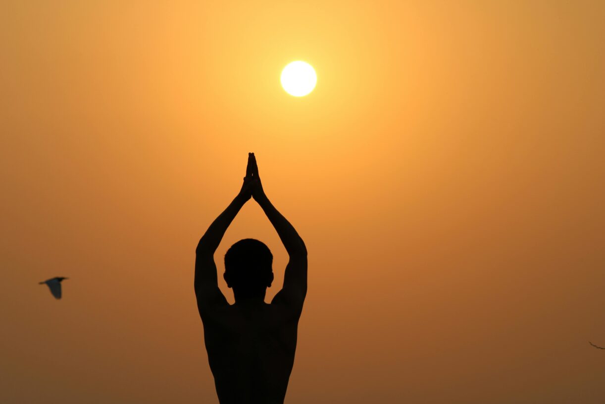 A serene silhouette of a person meditating with hands raised towards the sunset.
