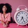 Pensive woman with afro hair leaning on a table next to a large pink alarm clock. 2026 में गृहिणियों के लिए टाइम मैनेजमेंट: जब 'मशीन' नहीं, 'मन' जीतता है समय की जंग