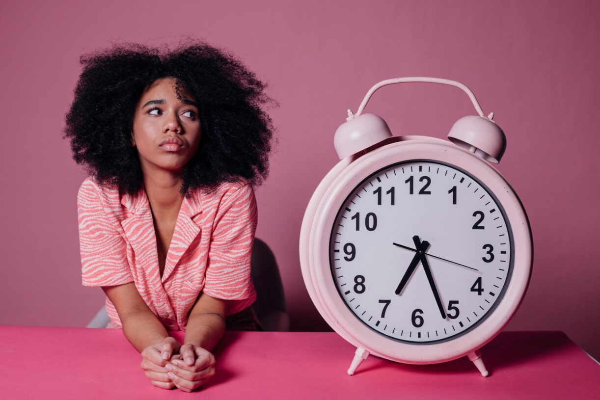 Pensive woman with afro hair leaning on a table next to a large pink alarm clock. 2026 में गृहिणियों के लिए टाइम मैनेजमेंट: जब 'मशीन' नहीं, 'मन' जीतता है समय की जंग