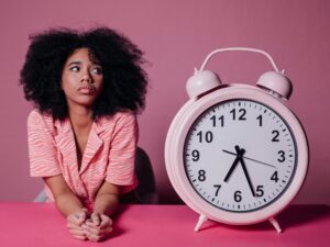 Pensive woman with afro hair leaning on a table next to a large pink alarm clock. 2026 में गृहिणियों के लिए टाइम मैनेजमेंट: जब 'मशीन' नहीं, 'मन' जीतता है समय की जंग